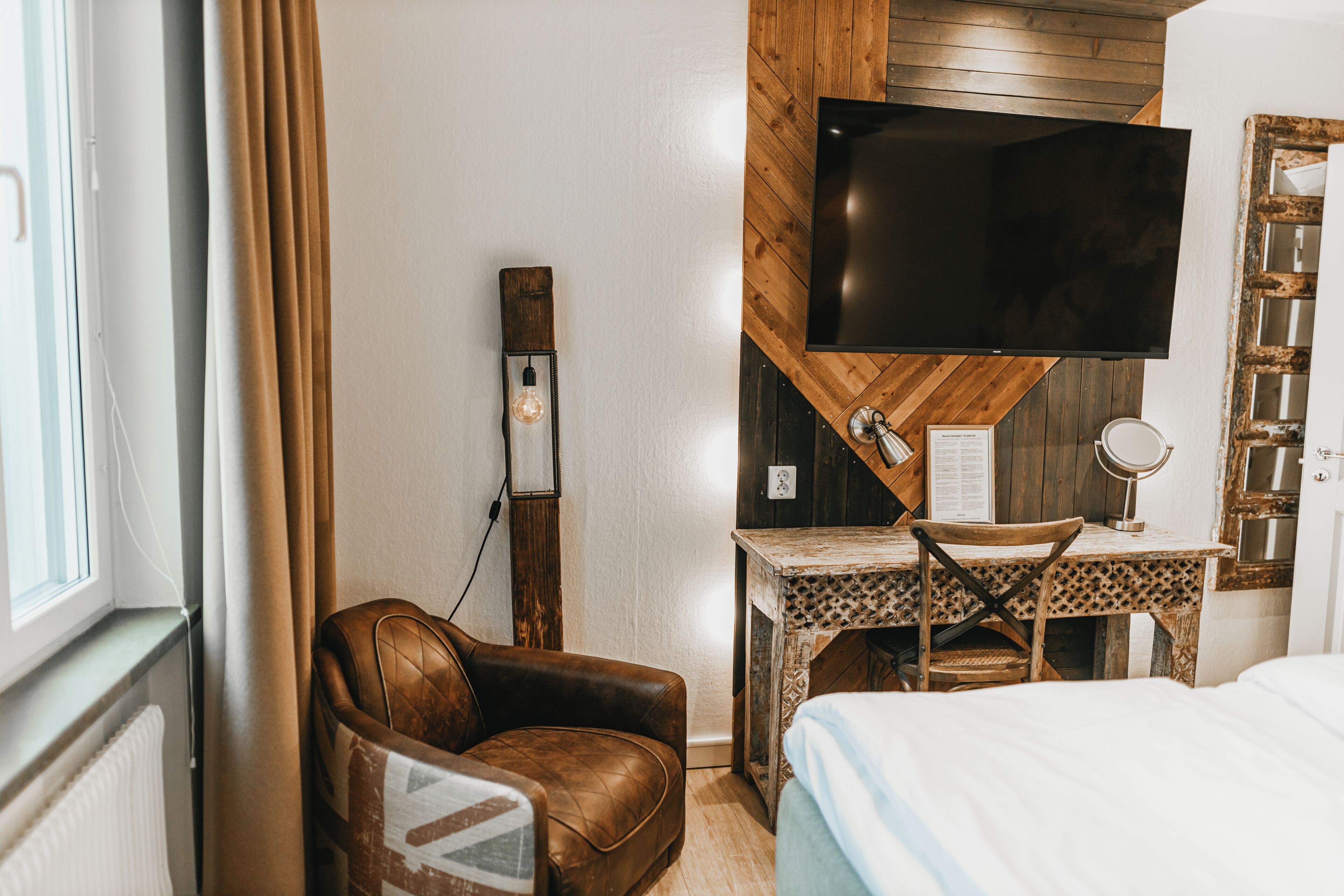 A hotel room with a bed, a desk with a chair, a television mounted on the wall, and a window with curtains.