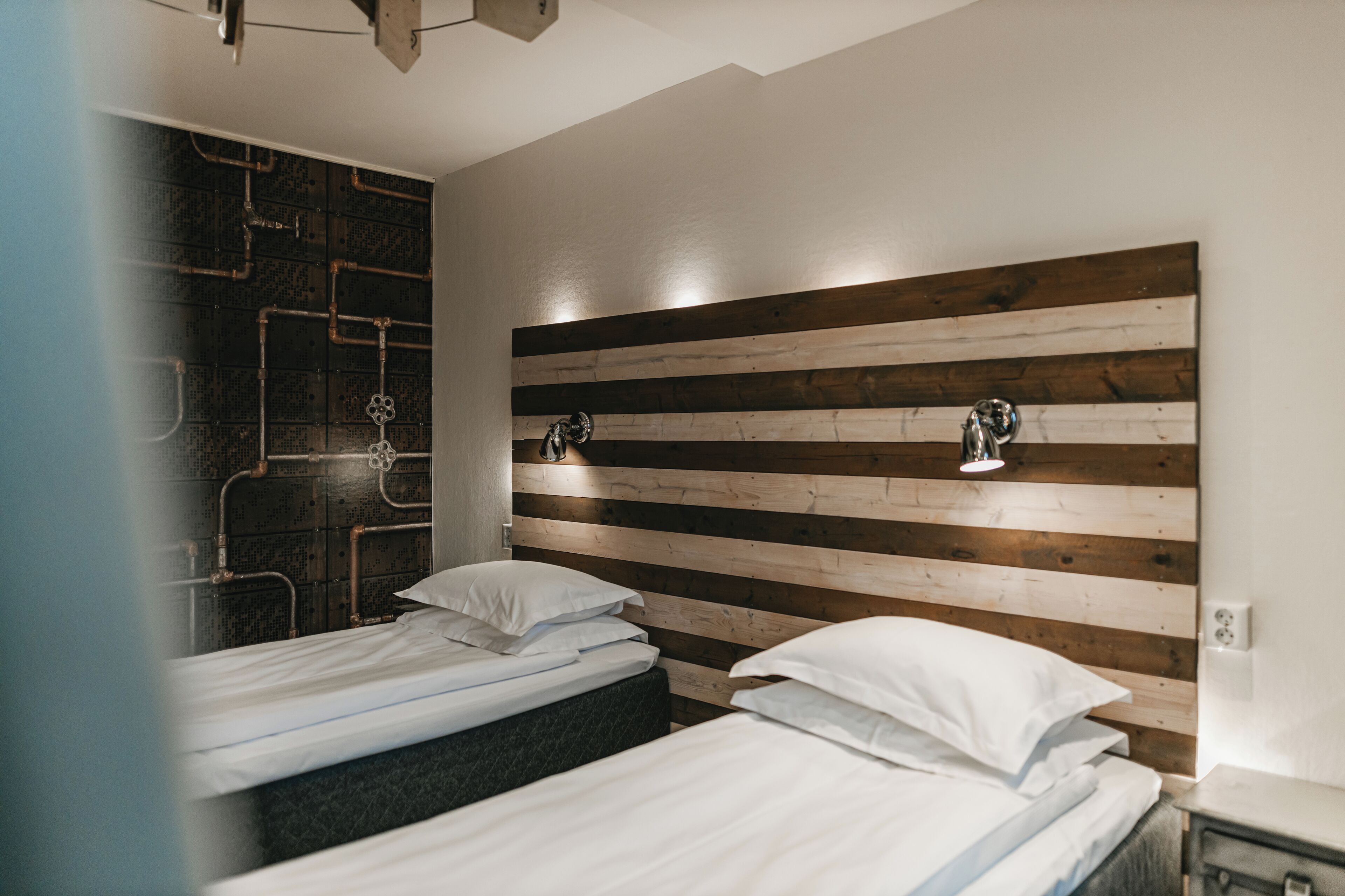 Une chambre d’hôtel moderne avec deux lits, une tête de lit en bois et un décor mural original composé de tuyaux.