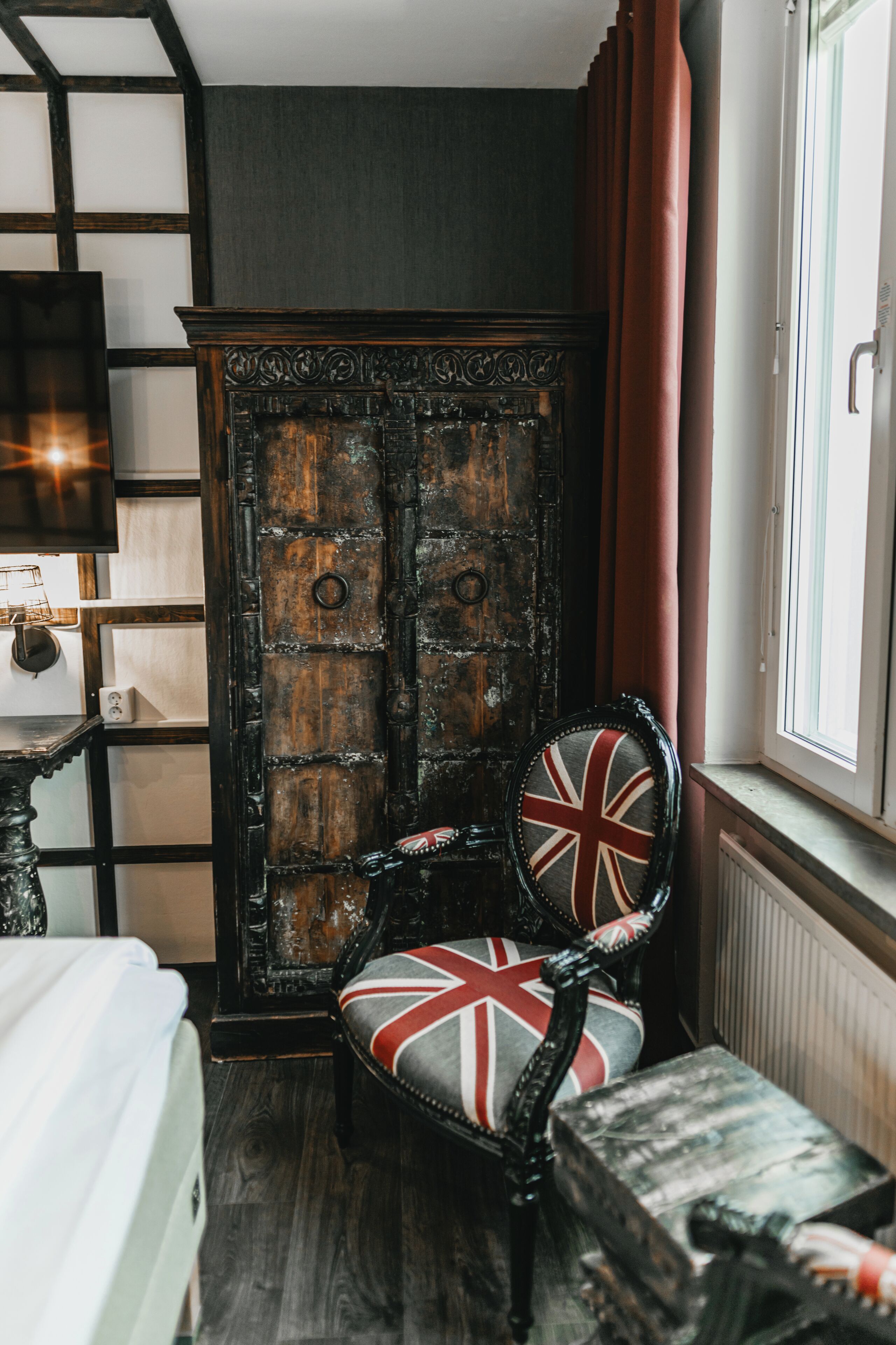 A room with a large wooden wardrobe, a chair with a Union Jack design, a bed, and a window with curtains.