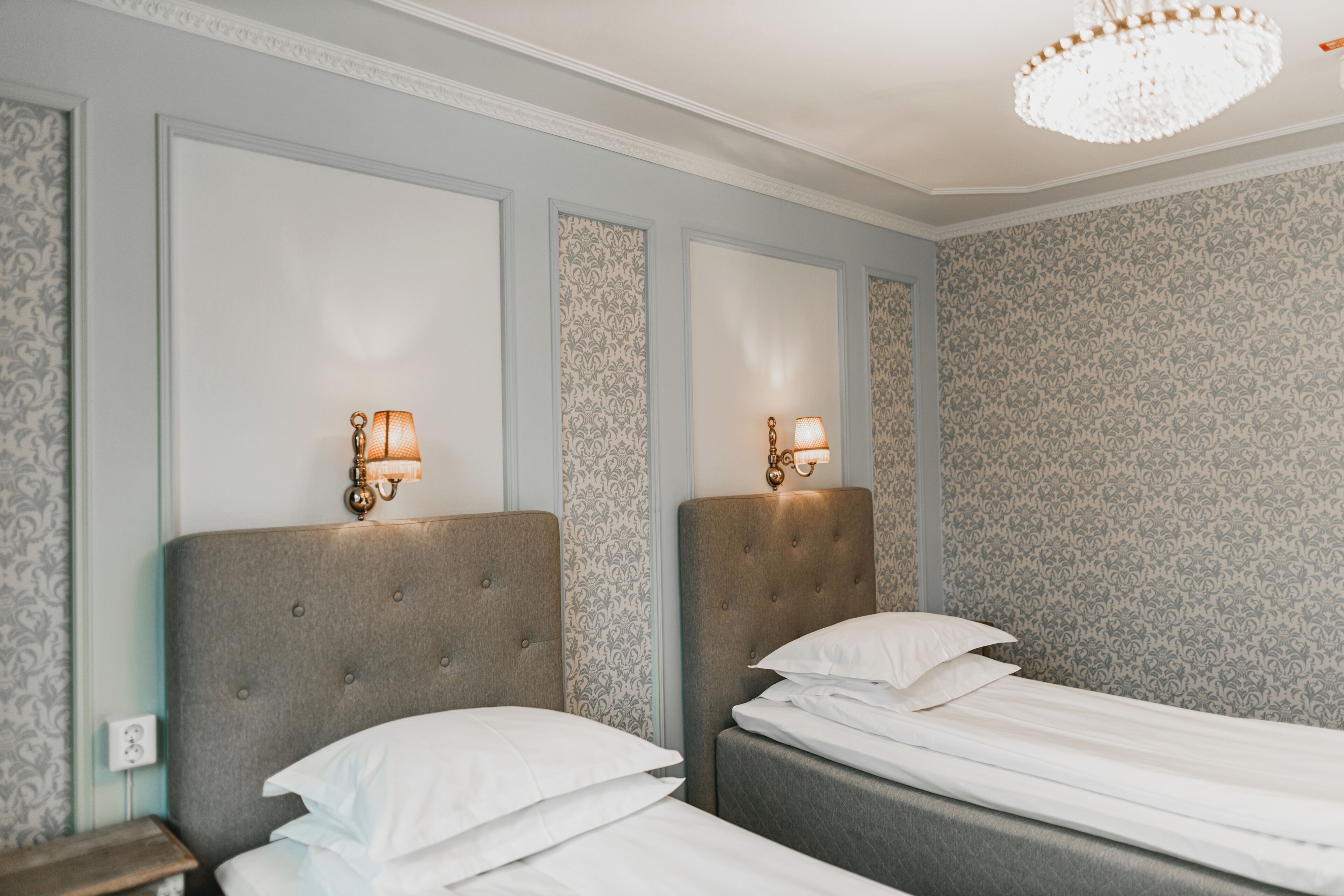 A hotel room with two beds, each with a headboard and a wall-mounted lamp.