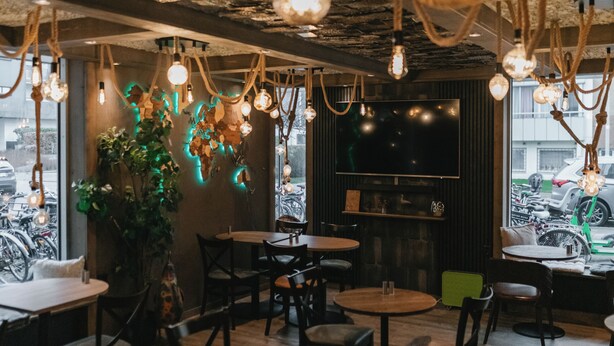 Ein Restaurantinneres mit Holztischen und -stühlen, hängenden Lampen und einem großen Fenster, vor dem Fahrräder stehen.