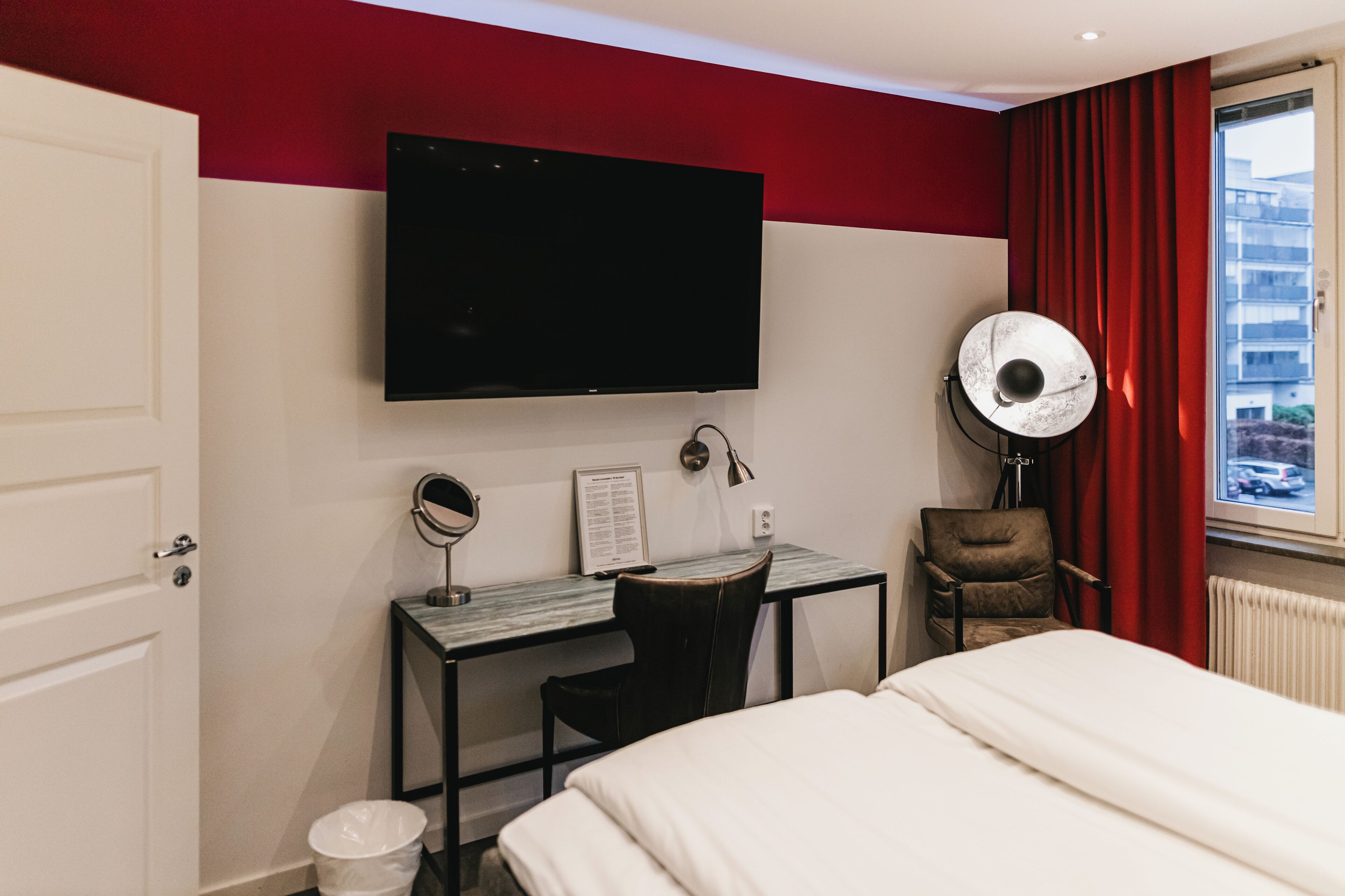 Habitación de hotel con cama, escritorio con silla, televisor de pantalla plana montado en la pared, espejo y ventana con cortinas rojas.
