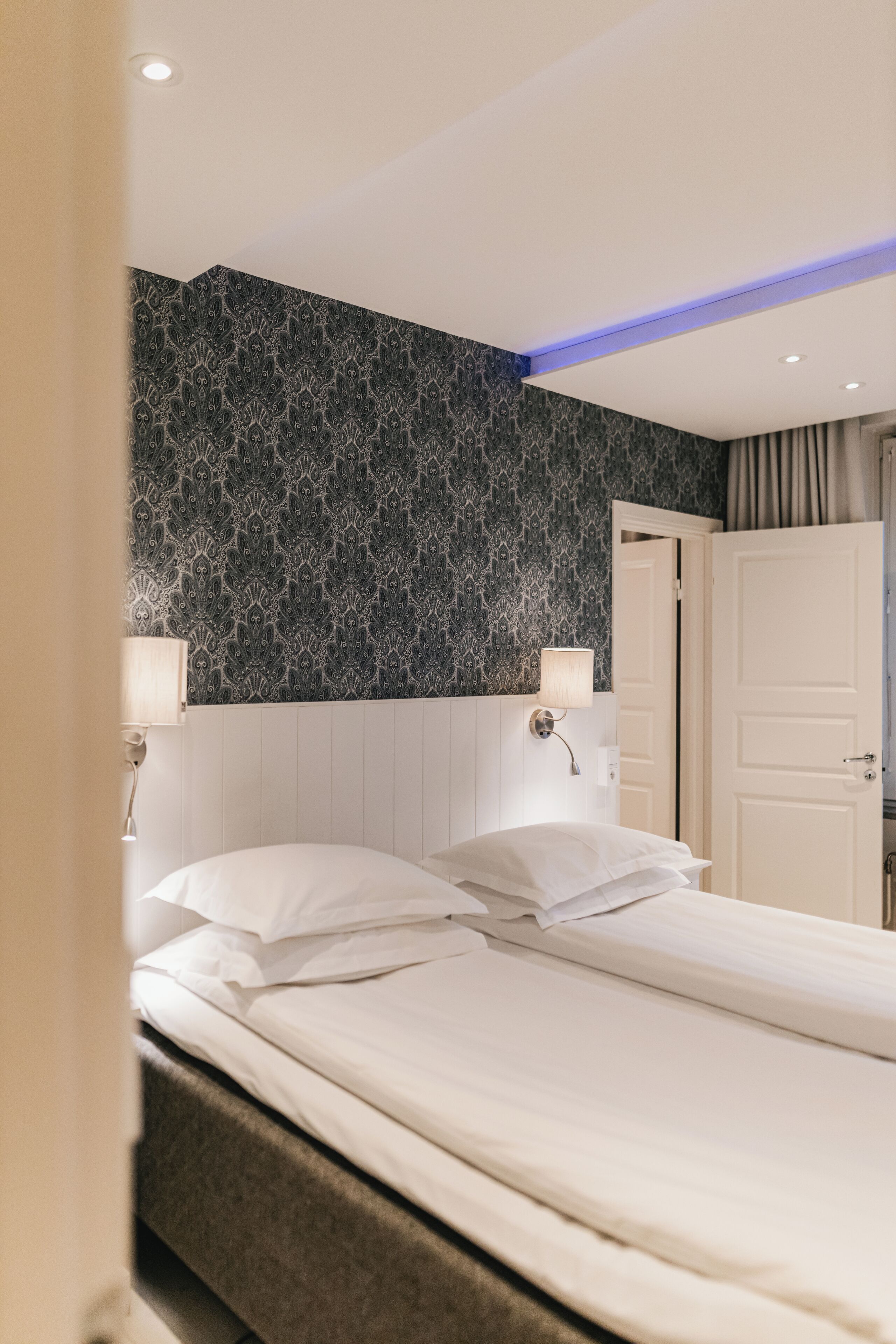 Habitación de hotel con cama, mesita de noche con lámpara, pared con estampado y una puerta.