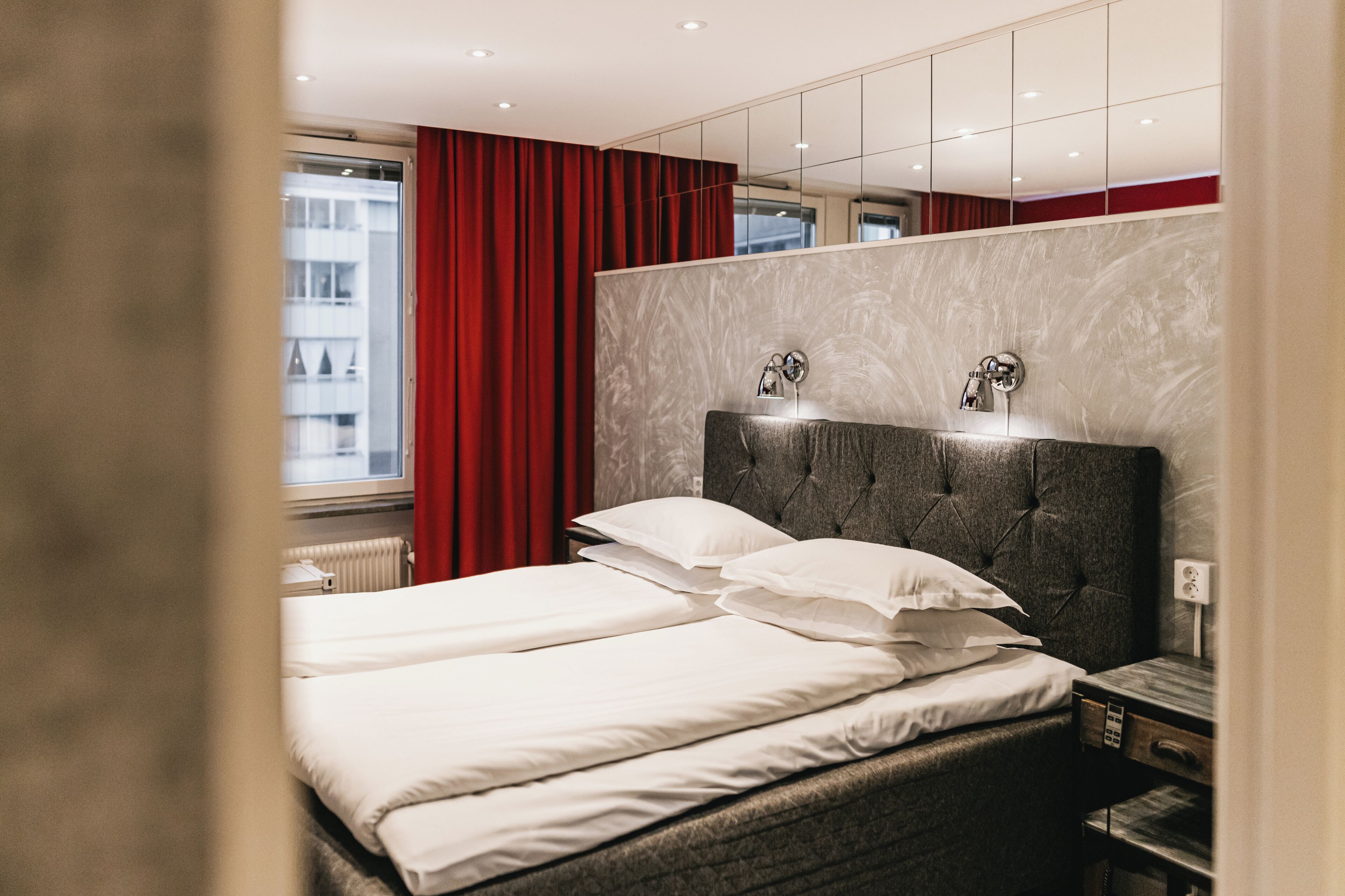 Hotellihuone, jossa on sänky, yöpöytä, peili ja ikkuna, jossa on punaiset verhot.