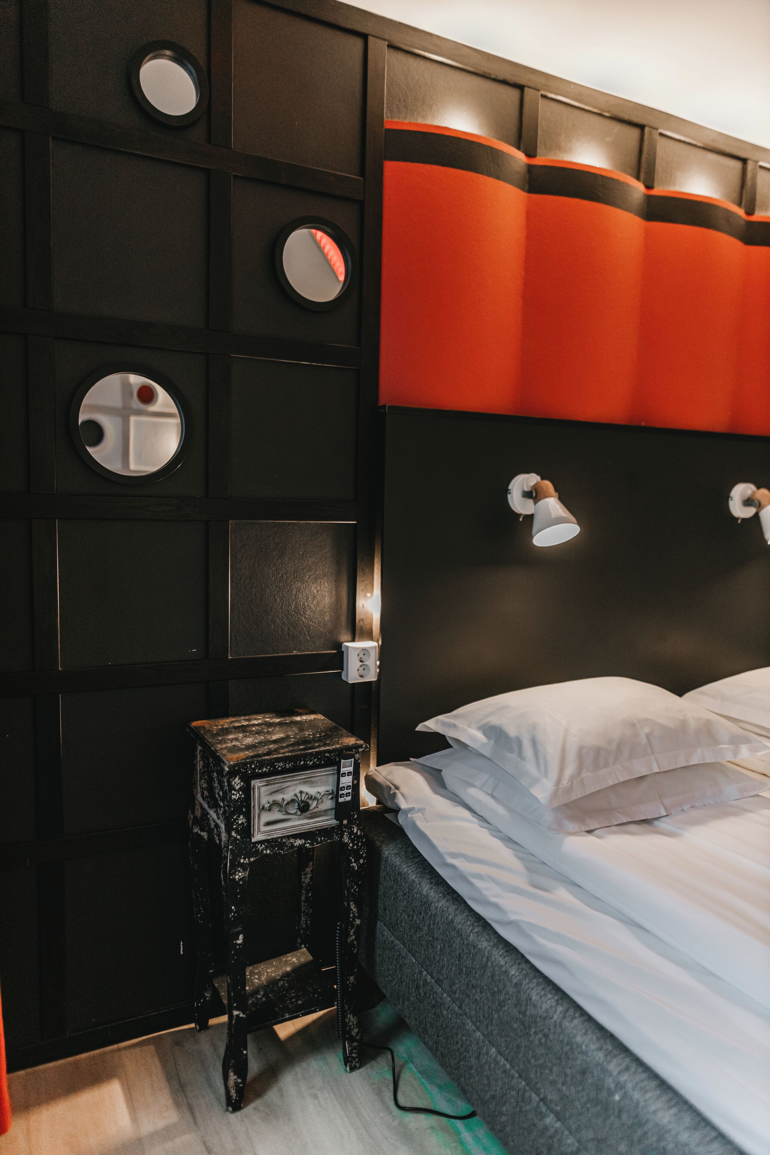 Quarto de hotel com cabeceira preta e laranja, uma pequena mesa de cabeceira e uma cama com roupa de cama branca.