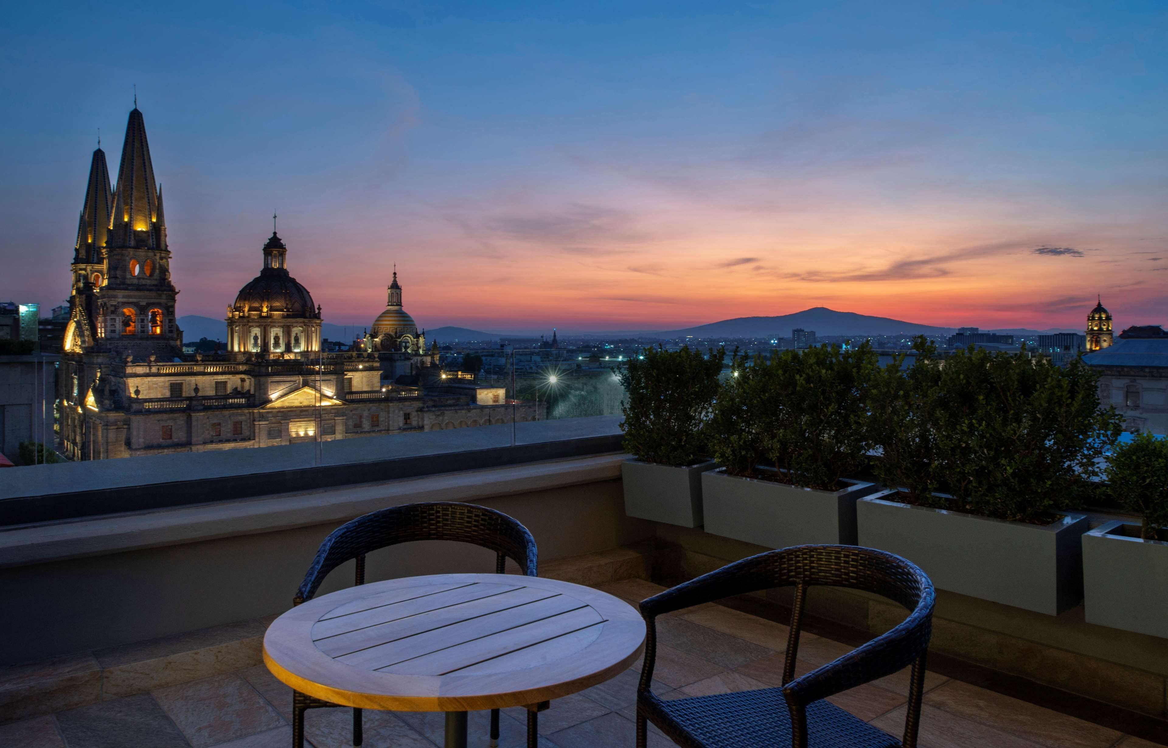 Une terrasse avec une table et des chaises donnant sur une vue panoramique de la ville au crépuscule, avec des bâtiments imposants et un coucher de soleil en toile de fond.