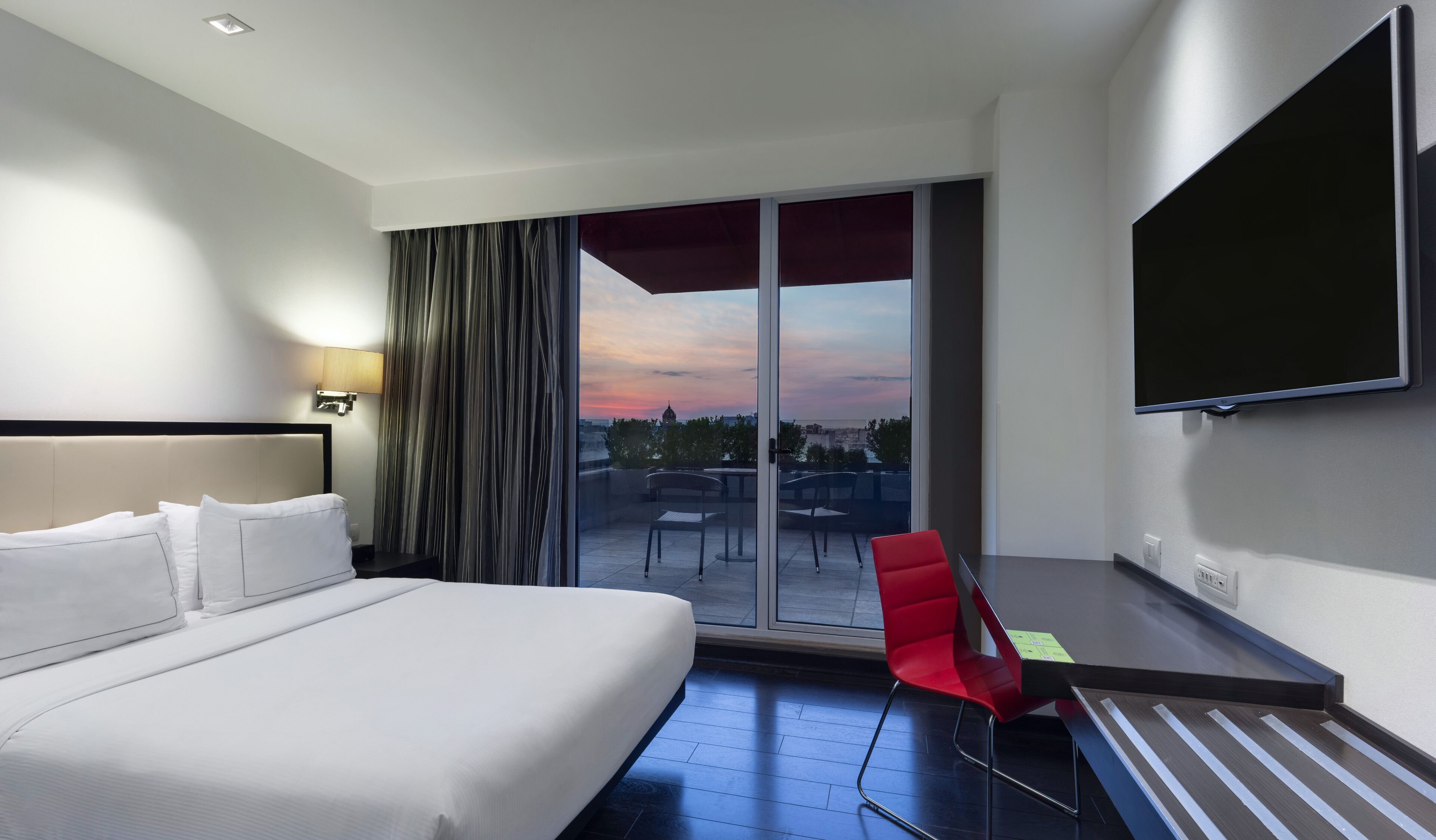 A hotel room with a bed, a desk with a chair, a TV, and a view of the cityscape through large windows.