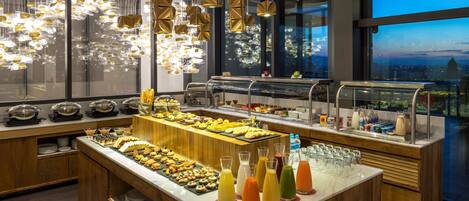 Montaje de bufé con diversos alimentos y bebidas, un ventanal con vistas a la ciudad e iluminación colgante moderna.