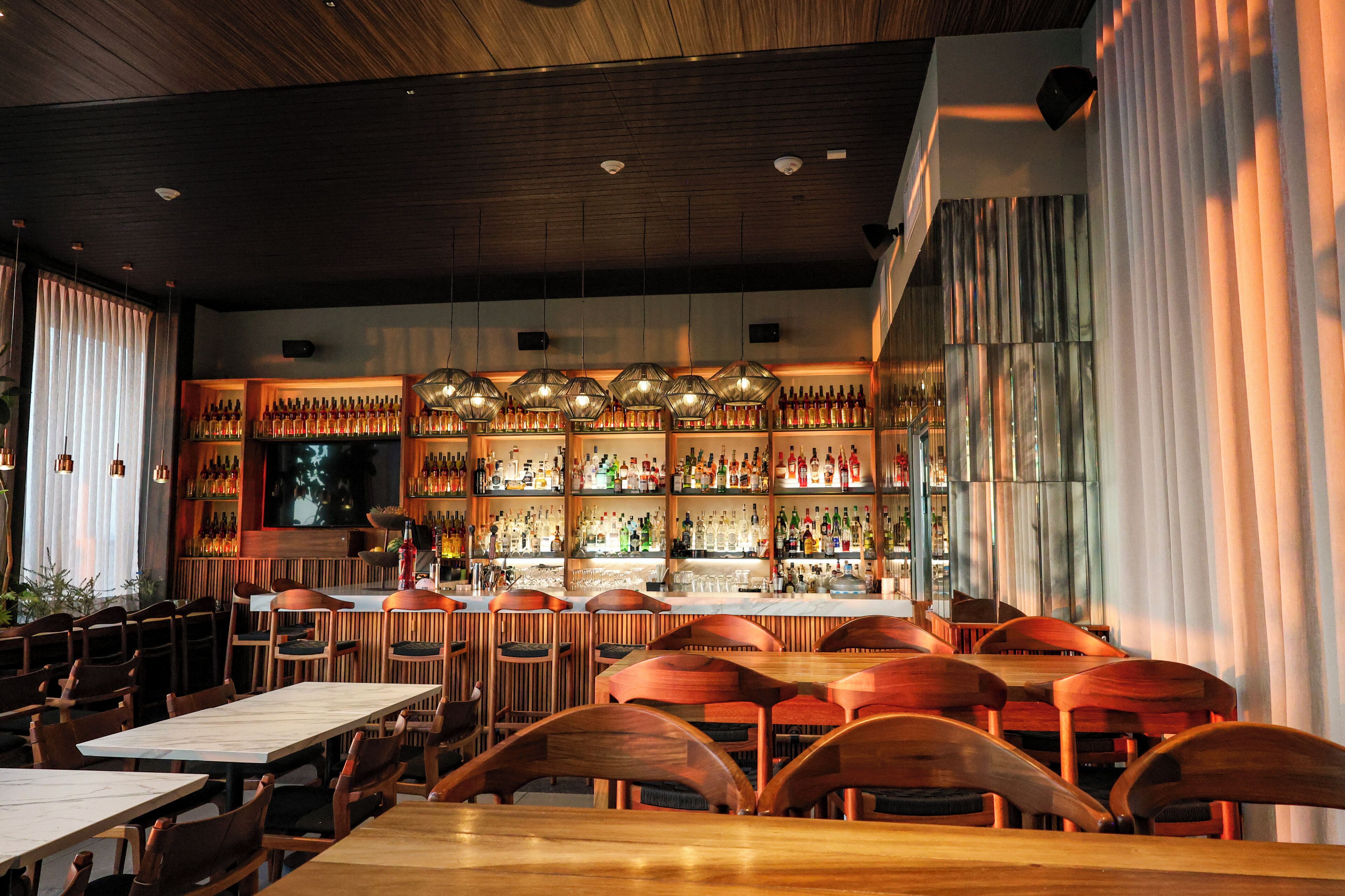 Interior de restaurante con mesas y sillas de madera, zona de bar con estanterías llenas de botellas e iluminación colgante.
