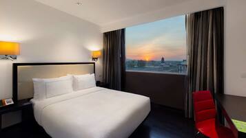 Habitación de hotel con una cama grande, una silla roja y vistas al atardecer sobre un paisaje urbano.