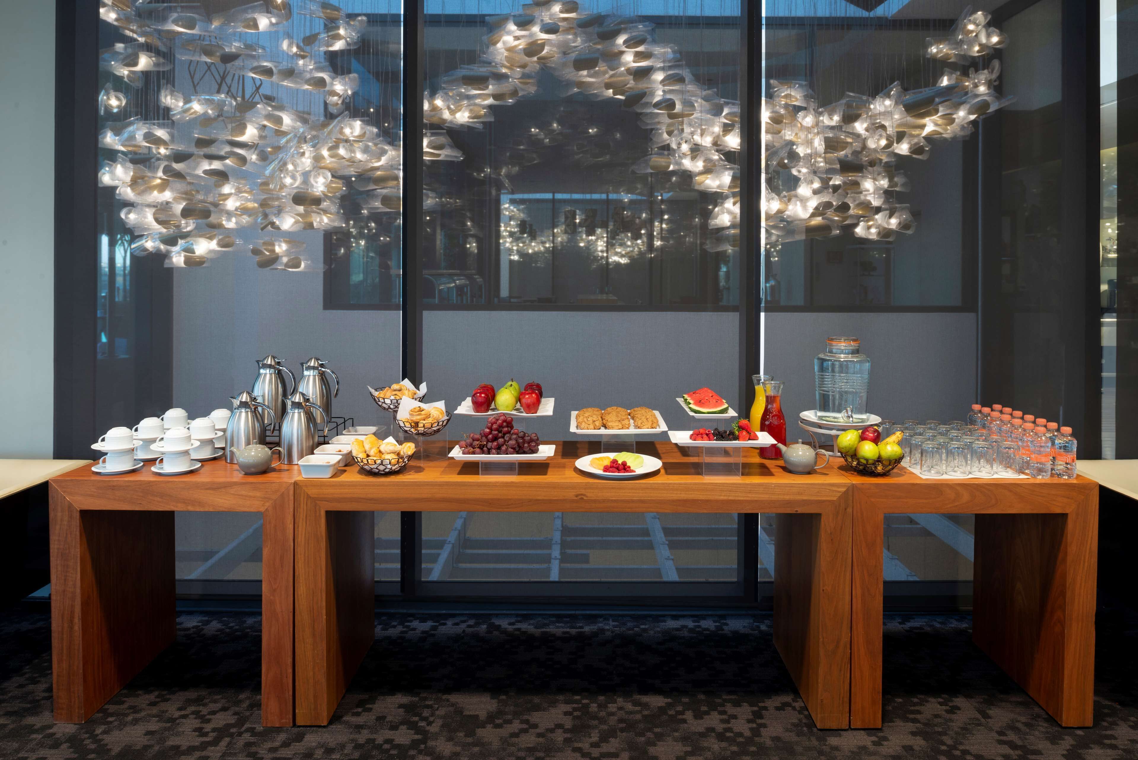 A breakfast buffet table with various food items and beverages, set against a modern interior with large windows and decorative lighting.