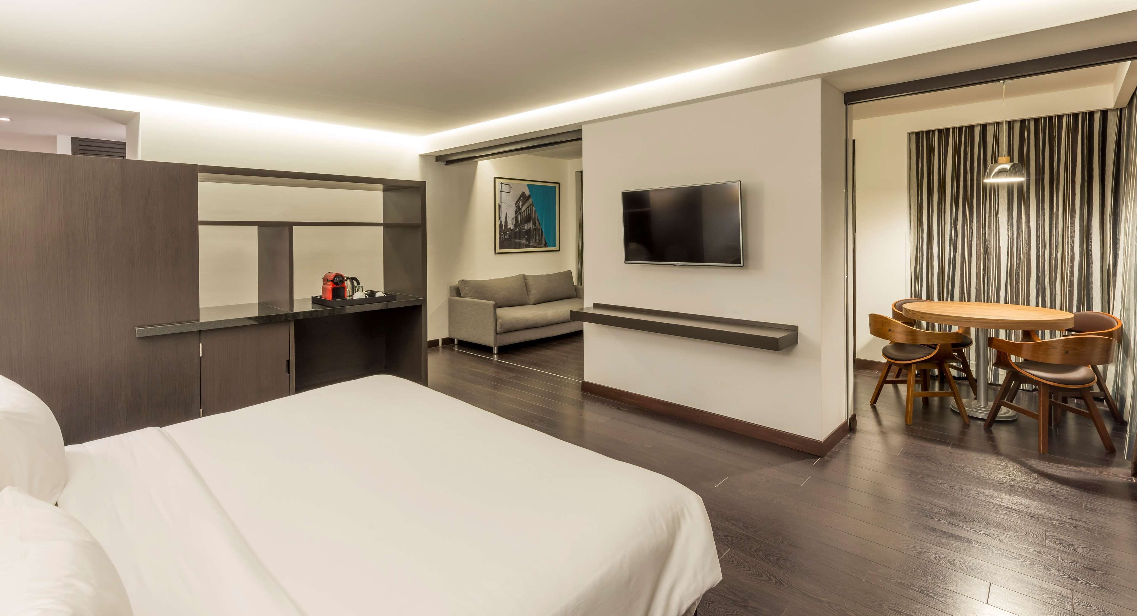 Una habitación de hotel moderna con una cama grande, un televisor de pantalla plana, un sofá, una zona de comedor con mesa y sillas, y un espejo grande.
