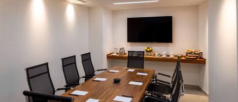 A conference room with a long wooden table, black chairs, a flat-screen TV, and a shelf with decorative items.
