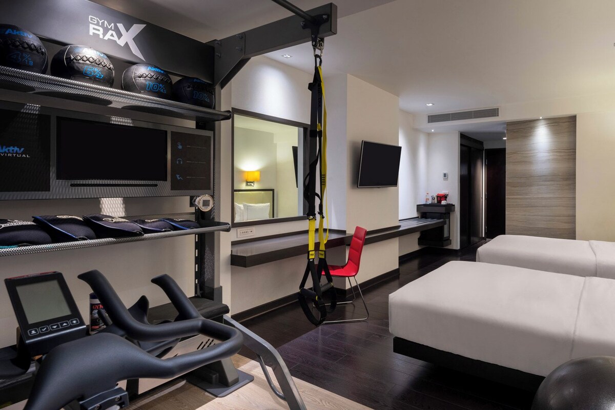 Une chambre d’hôtel moderne équipée d’un tapis de course, de ballons d’exercice, d’un fauteuil rouge, d’un lit et d’un téléviseur.