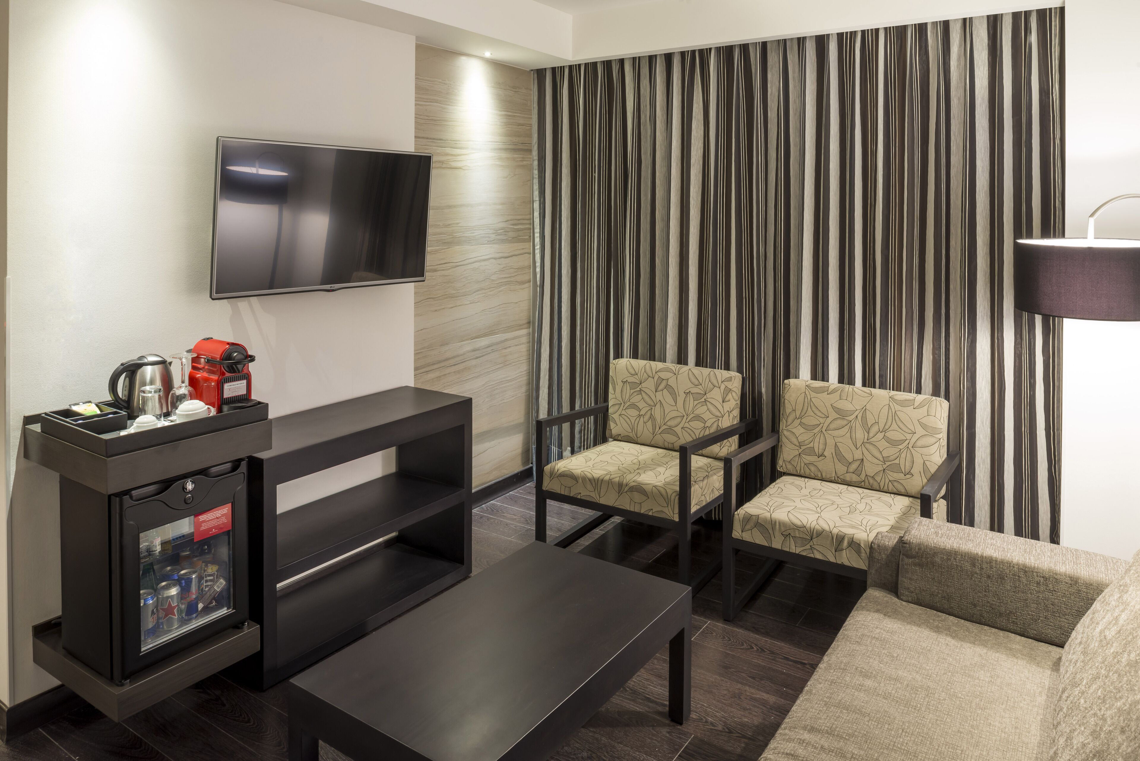 A modern living room with a flat-screen TV, a coffee machine, a small refrigerator, and two patterned armchairs.