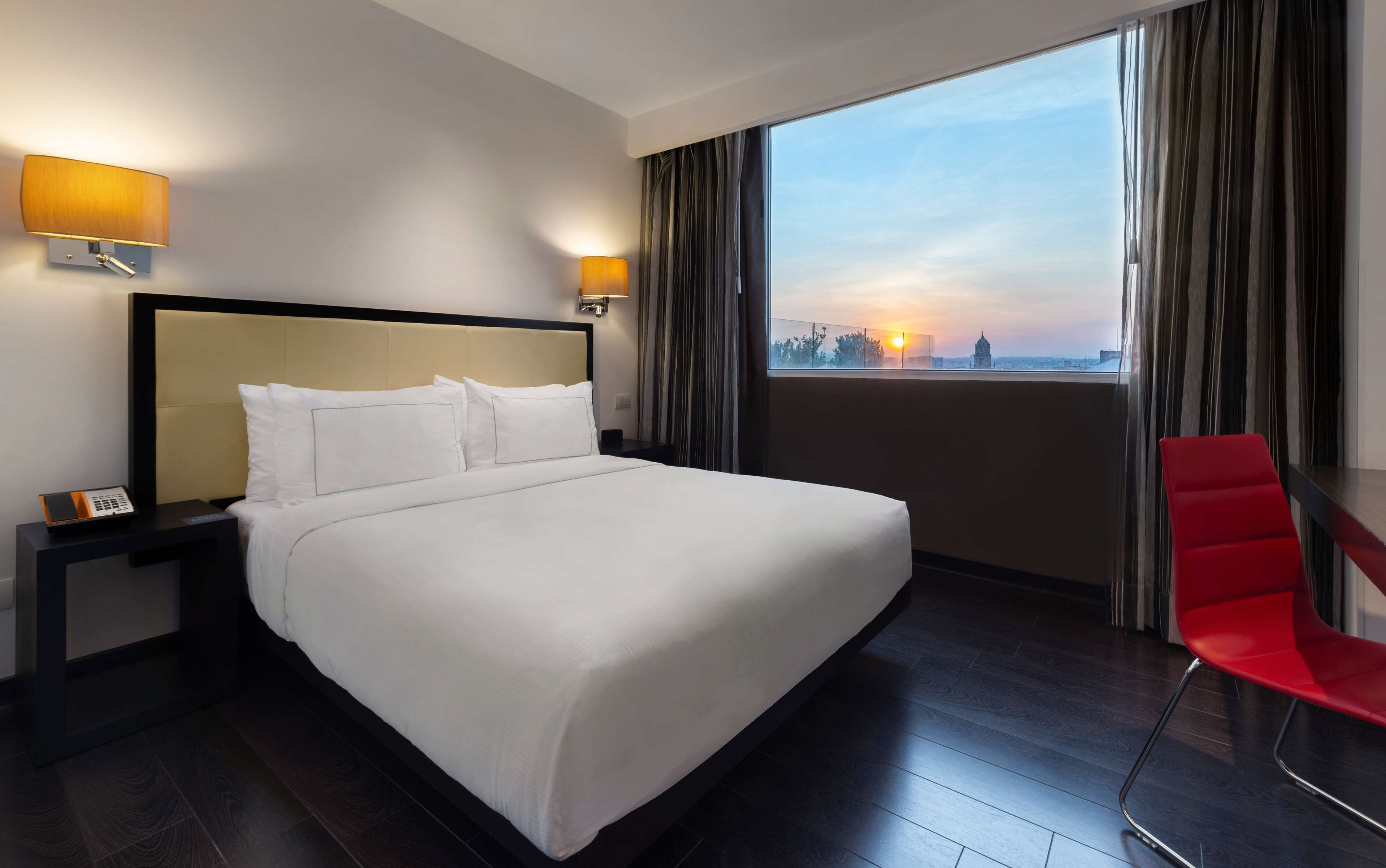 Habitación de hotel con una cama grande, dos lámparas de noche, una silla roja y vistas a la ciudad al atardecer.