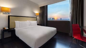 A hotel room with a large bed, two bedside lamps, a red chair, and a view of the cityscape at sunset.