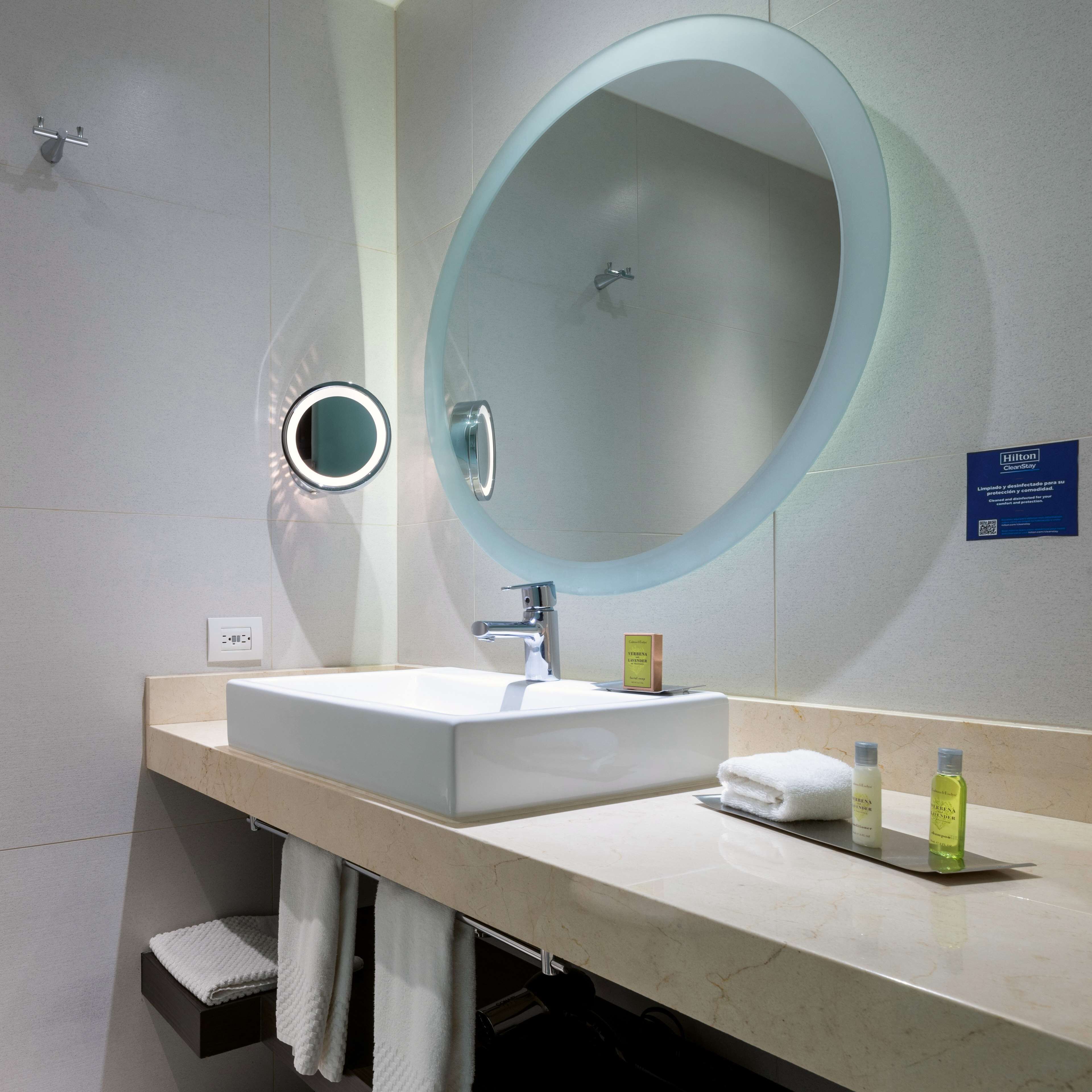 A modern bathroom with a large oval mirror, a white sink, and two toiletries on the counter.