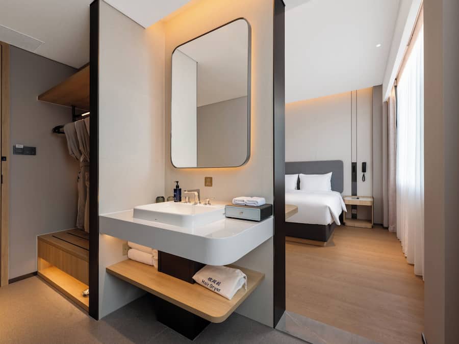 Una habitación de hotel moderna con un lavamanos grande en el baño, un espejo, una mesita de noche, una cama y un pequeño banco de madera.