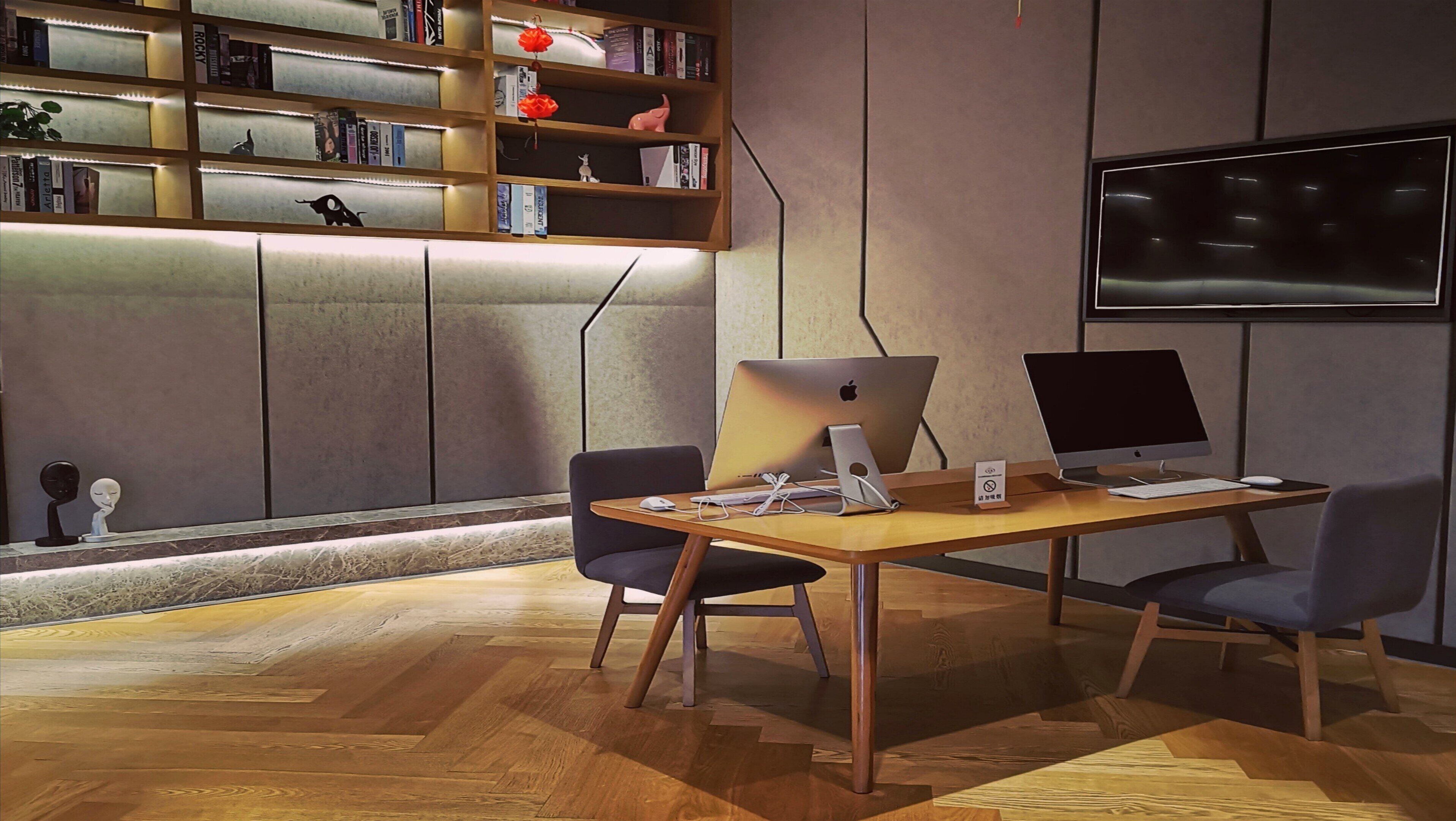Ein moderner Büroarbeitsplatz mit zwei Schreibtischen, jeweils ausgestattet mit einem Apple iMac, einem Laptop und einer Maus. Bücherregale mit diversen Gegenständen und ein an der Wand befestigtes Fernsehgerät sind ebenfalls vorhanden.