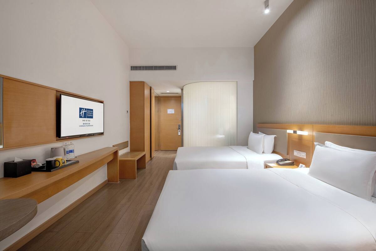 Habitación de hotel con dos camas, un banco de madera, un escritorio con teléfono y un cartel enmarcado en la pared.