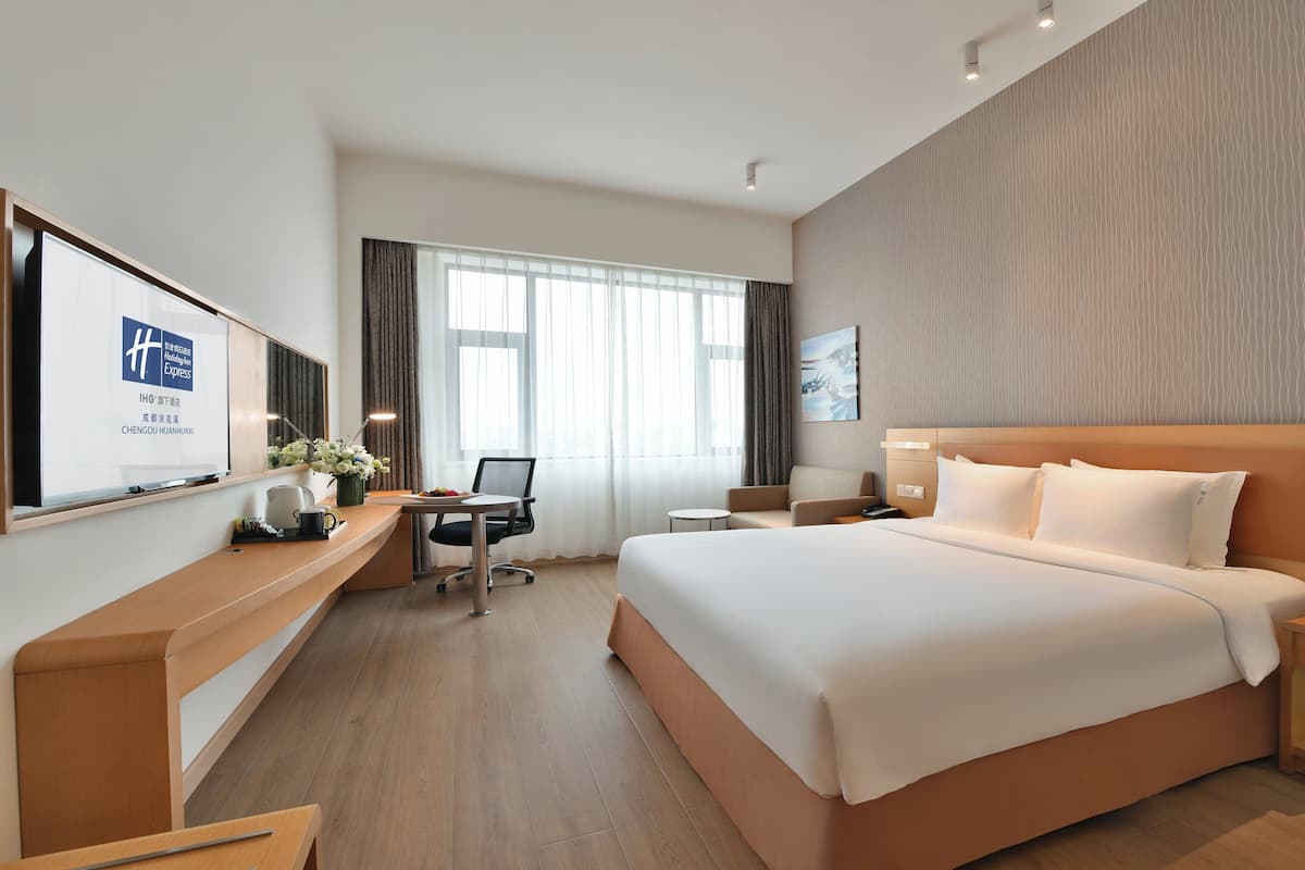 Habitación de hotel con una cama grande, un escritorio con computadora, una silla y una mesita pequeña. Hay una ventana con cortinas, un televisor montado en la pared y un cuadro.