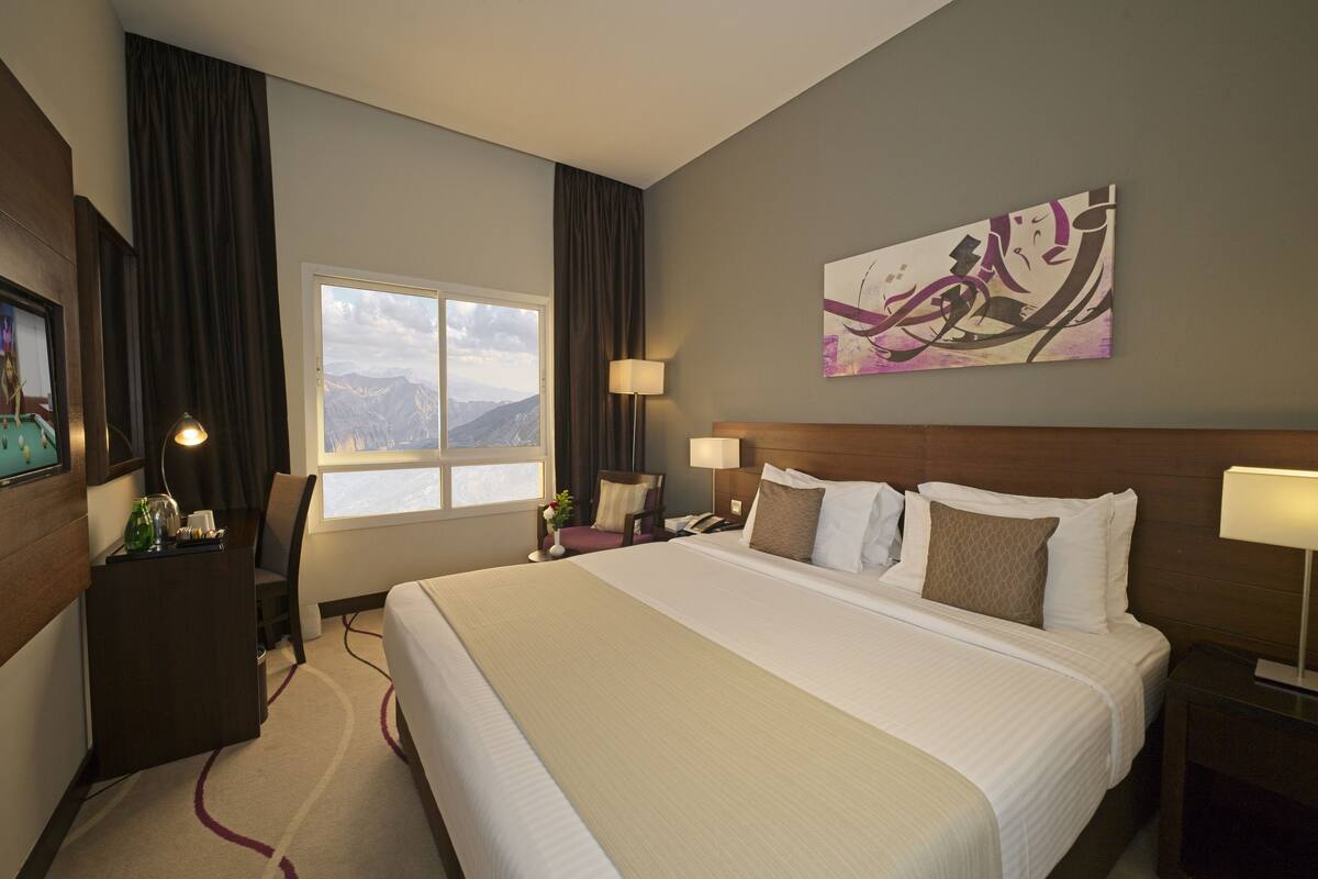 Ein Hotelzimmer mit einem großen Bett, einem Schreibtisch mit Lampe, einem Sessel, einem Nachttisch, einem Bild an der Wand und einem Fenster mit Bergblick.