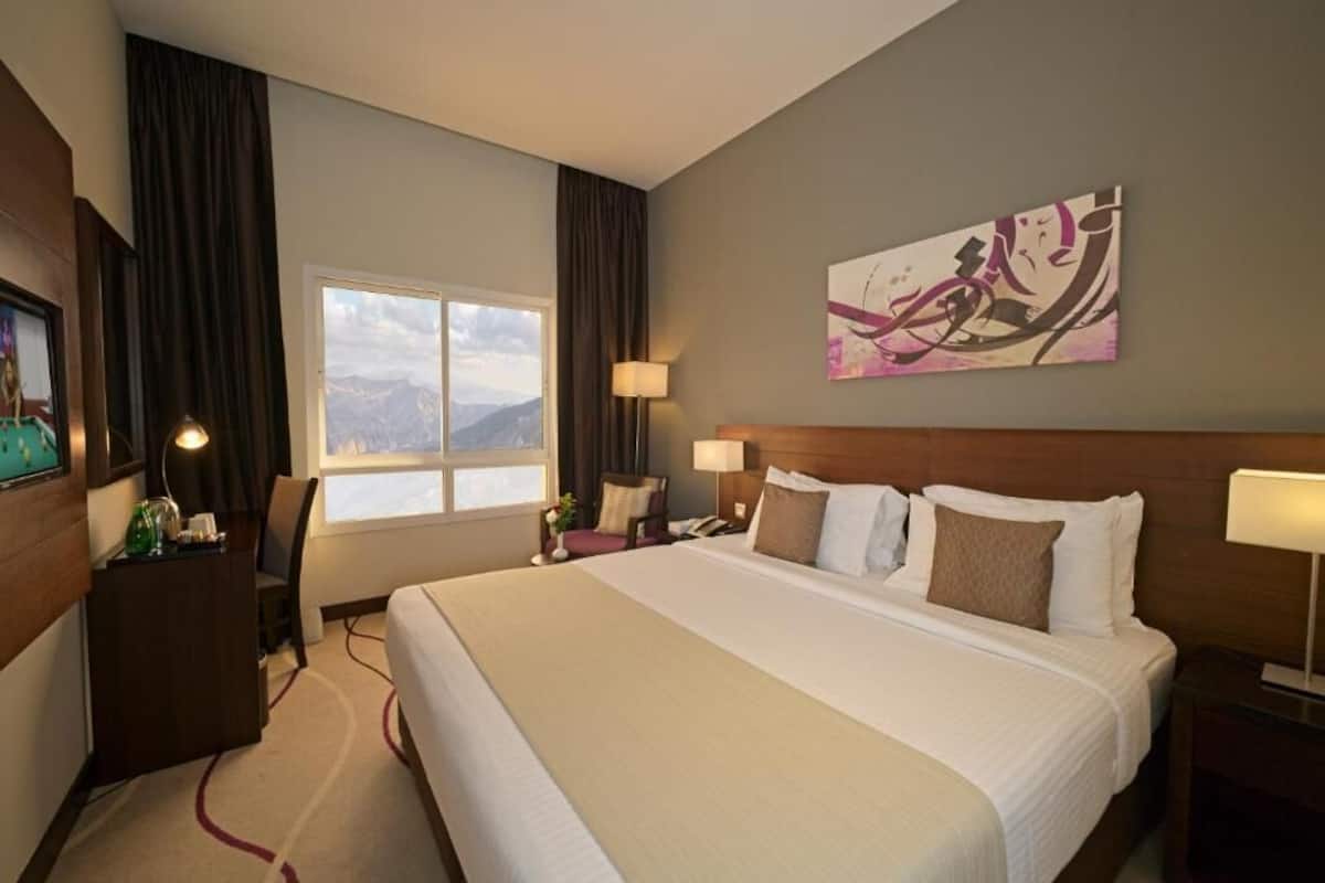 Ein Hotelzimmer mit einem großen Bett, einem Schreibtisch, einem Sessel, einem Nachttisch, einer Lampe, einem Bild an der Wand und einem Fenster mit Bergblick.