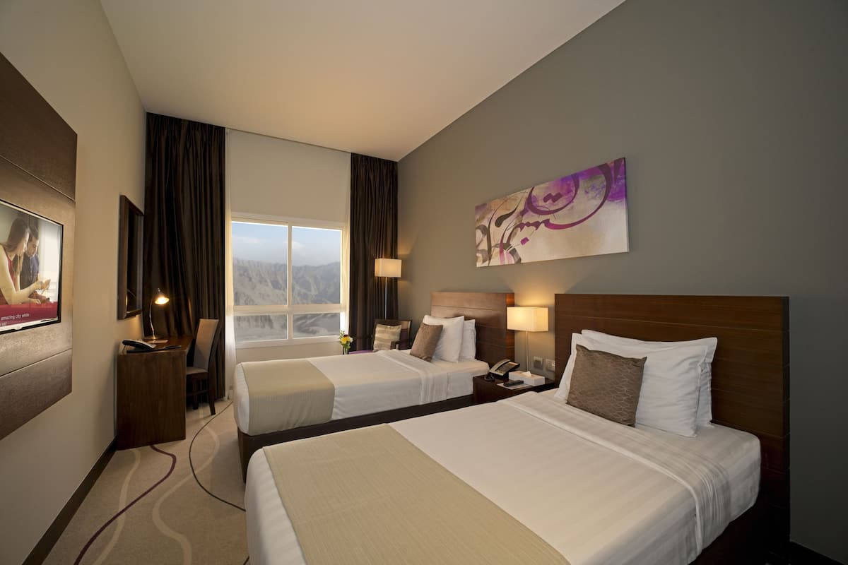 Ein Hotelzimmer mit zwei Betten, einem Schreibtisch, einem Stuhl, einem Fernseher und einem Fenster mit Bergblick.