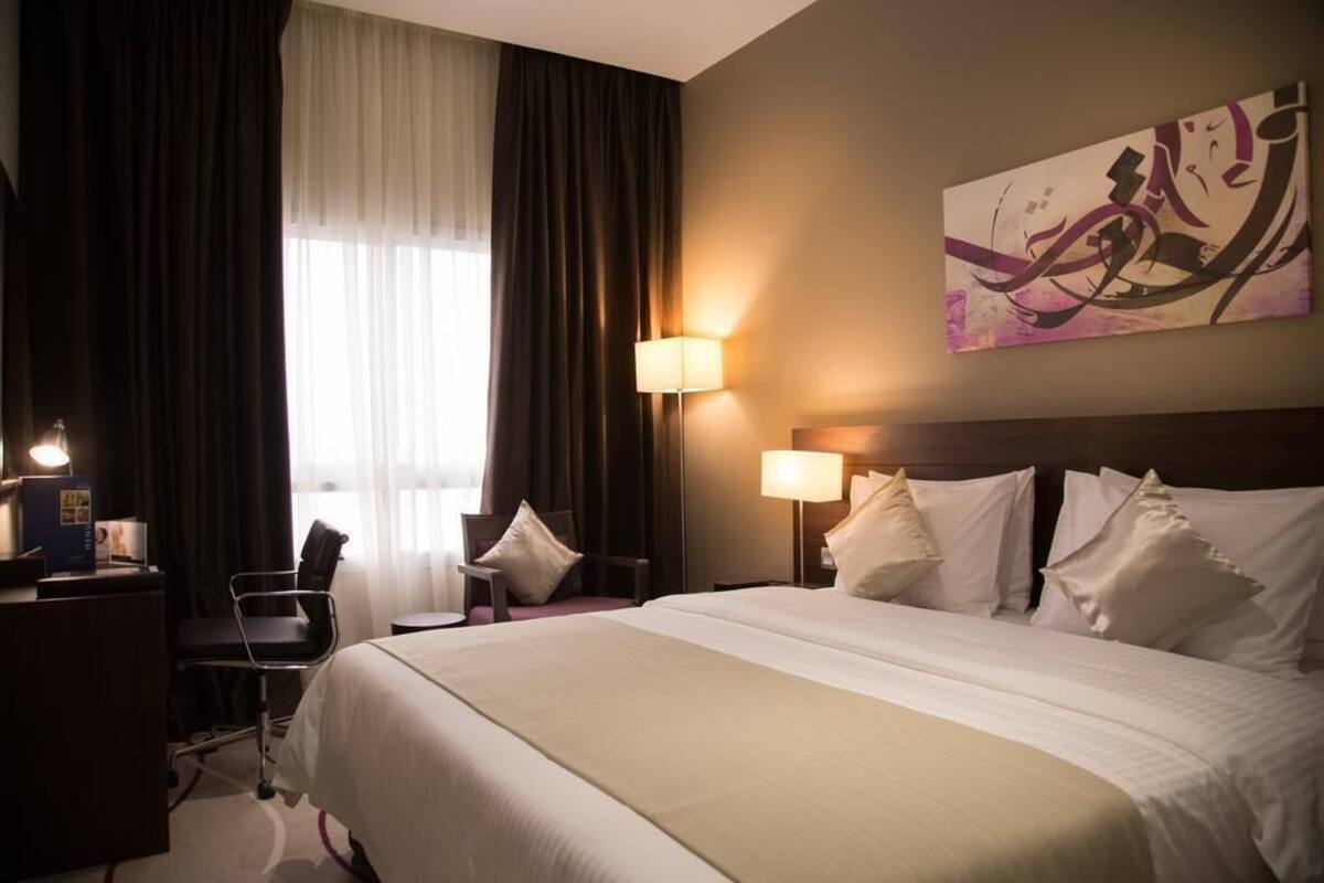 Une chambre d’hôtel avec un grand lit, deux lampes de chevet, un bureau avec une lampe, une chaise et une fenêtre avec des rideaux.