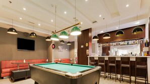 A pool table in a room with a bar area, red seating, and pendant lighting.
