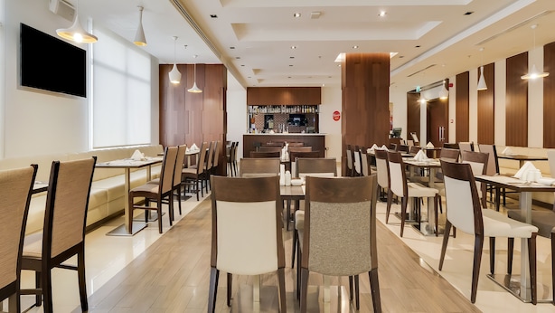 A modern restaurant interior with multiple dining tables and chairs, a bar area, and a flat-screen TV mounted on the wall.