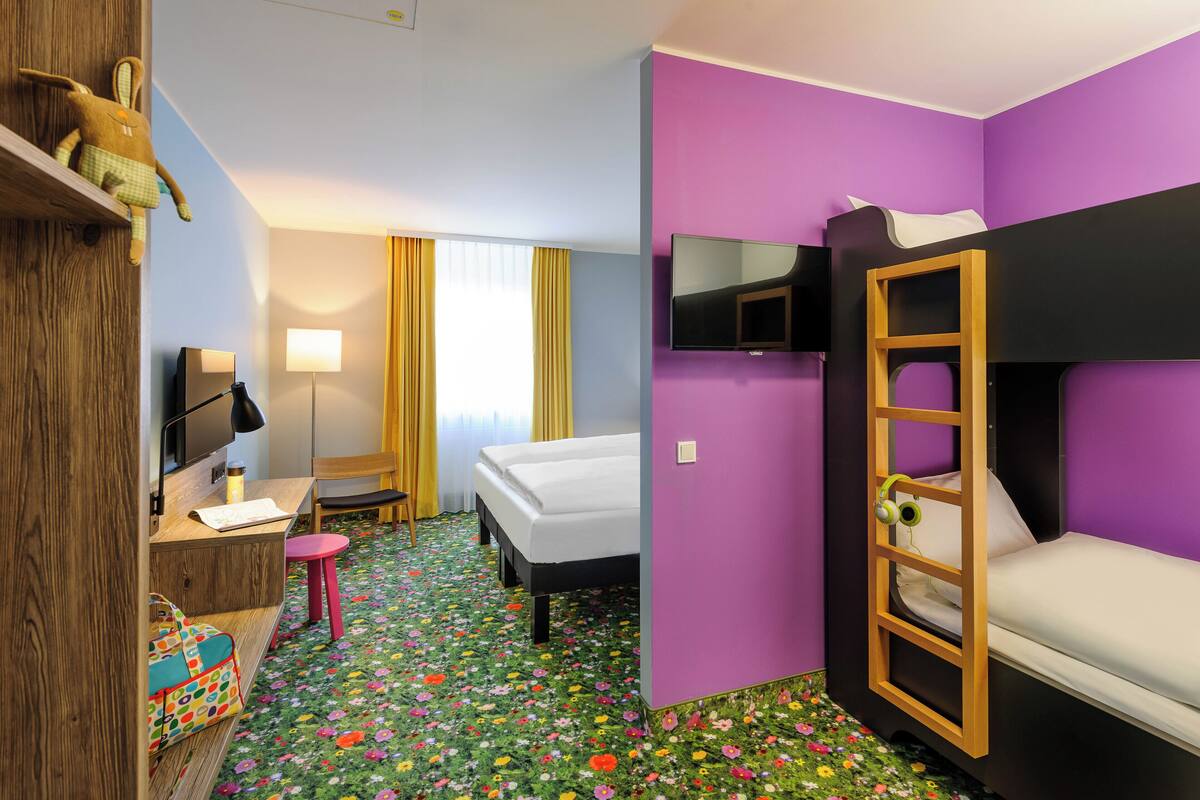 Une chambre avec un lit superposé, un bureau avec une lampe, une chaise, une petite table et un tapis à motifs floraux aux couleurs vives.