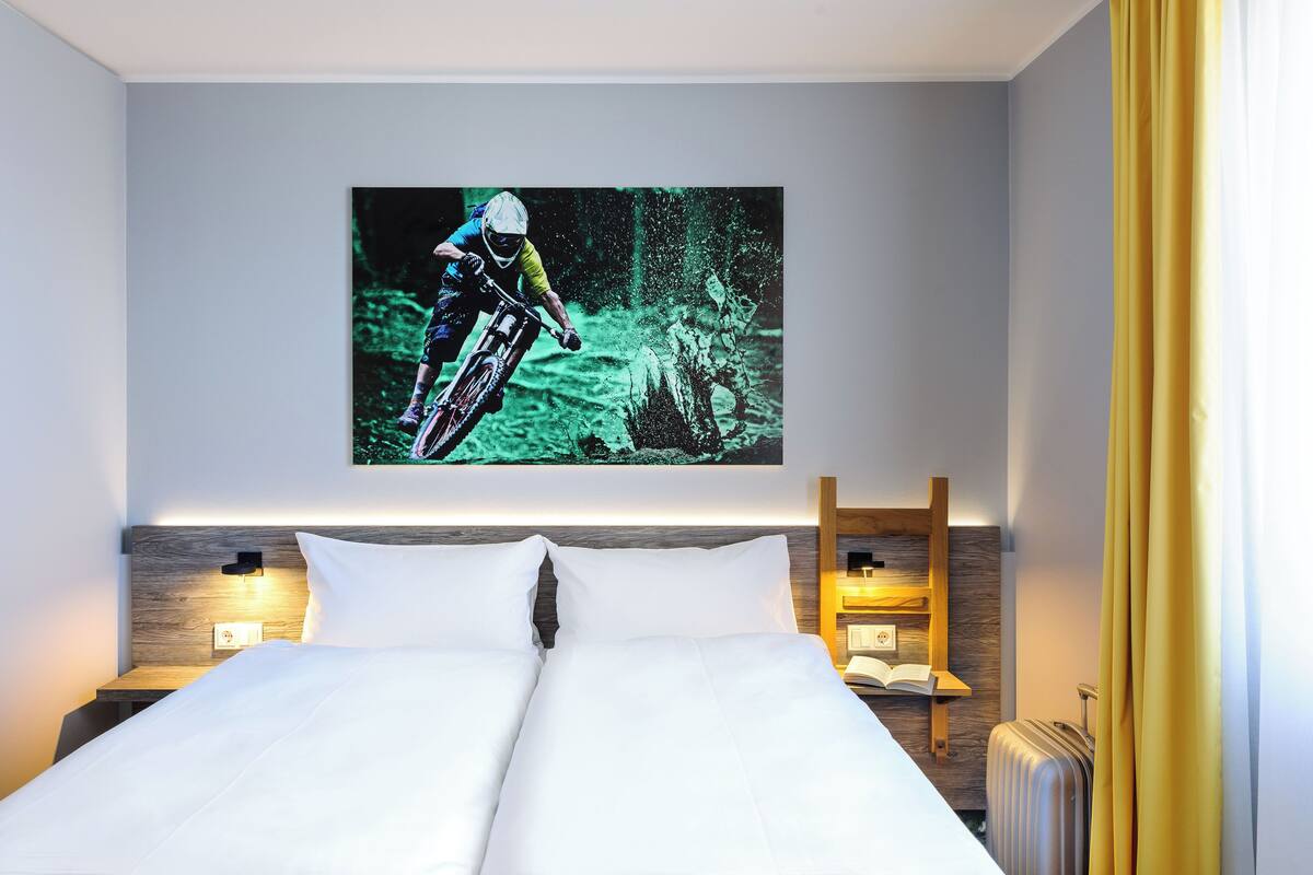Une chambre d’hôtel avec un lit, un téléviseur fixé au mur qui diffuse des images d’un vététiste, des lampes de chevet et une tête de lit en bois.