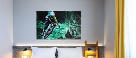 A hotel room with a bed, a mounted TV displaying a mountain biker, bedside lamps, and a wooden headboard.