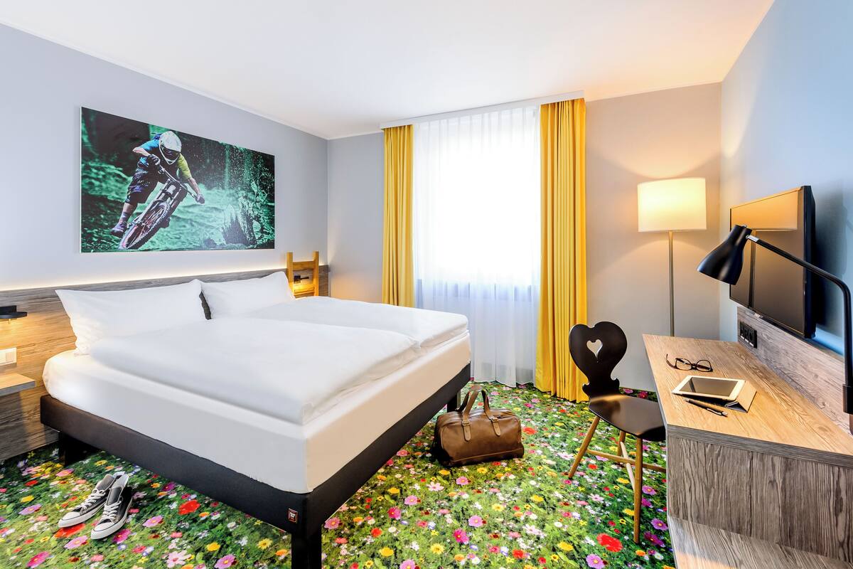 Une chambre d’hôtel avec un lit, un bureau avec un ordinateur portable, une chaise, une télévision et une grande fenêtre avec des rideaux.