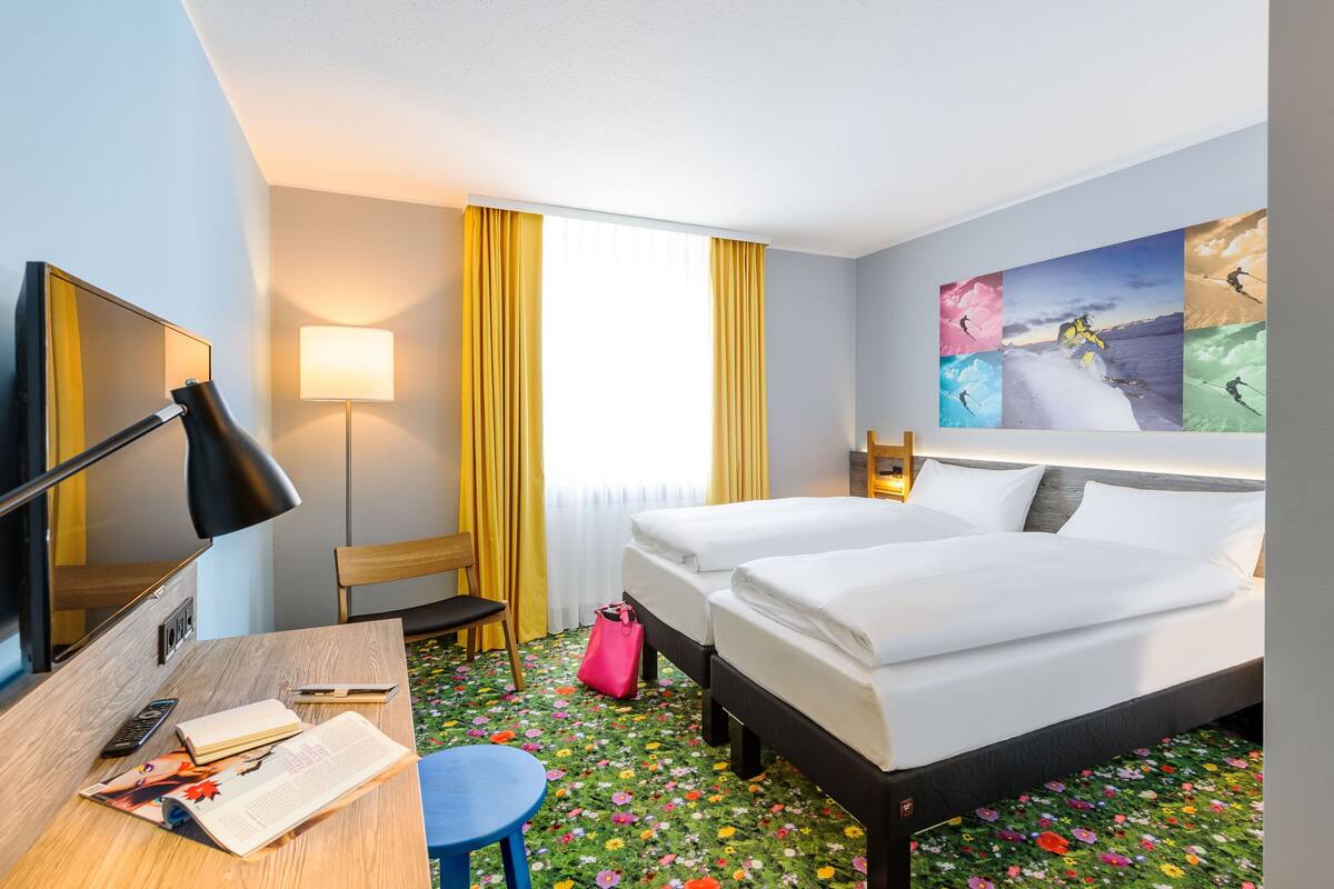 Une chambre d’hôtel avec deux lits, un bureau avec une lampe, une chaise, une petite table, une moquette colorée et une grande fenêtre avec des rideaux.