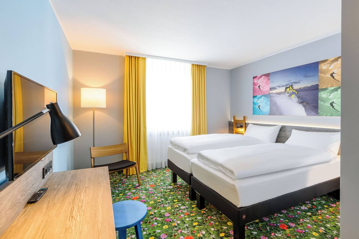 Une chambre d’hôtel avec deux lits, un bureau avec une lampe, une chaise, une télévision et une grande fenêtre avec des rideaux.