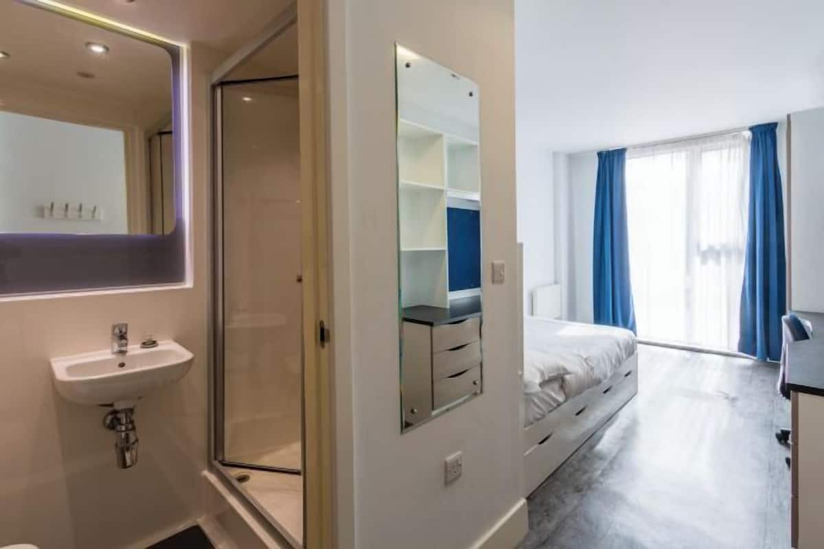 A hotel room with a bed, a bathroom with a sink, a mirror, and a window with curtains.
