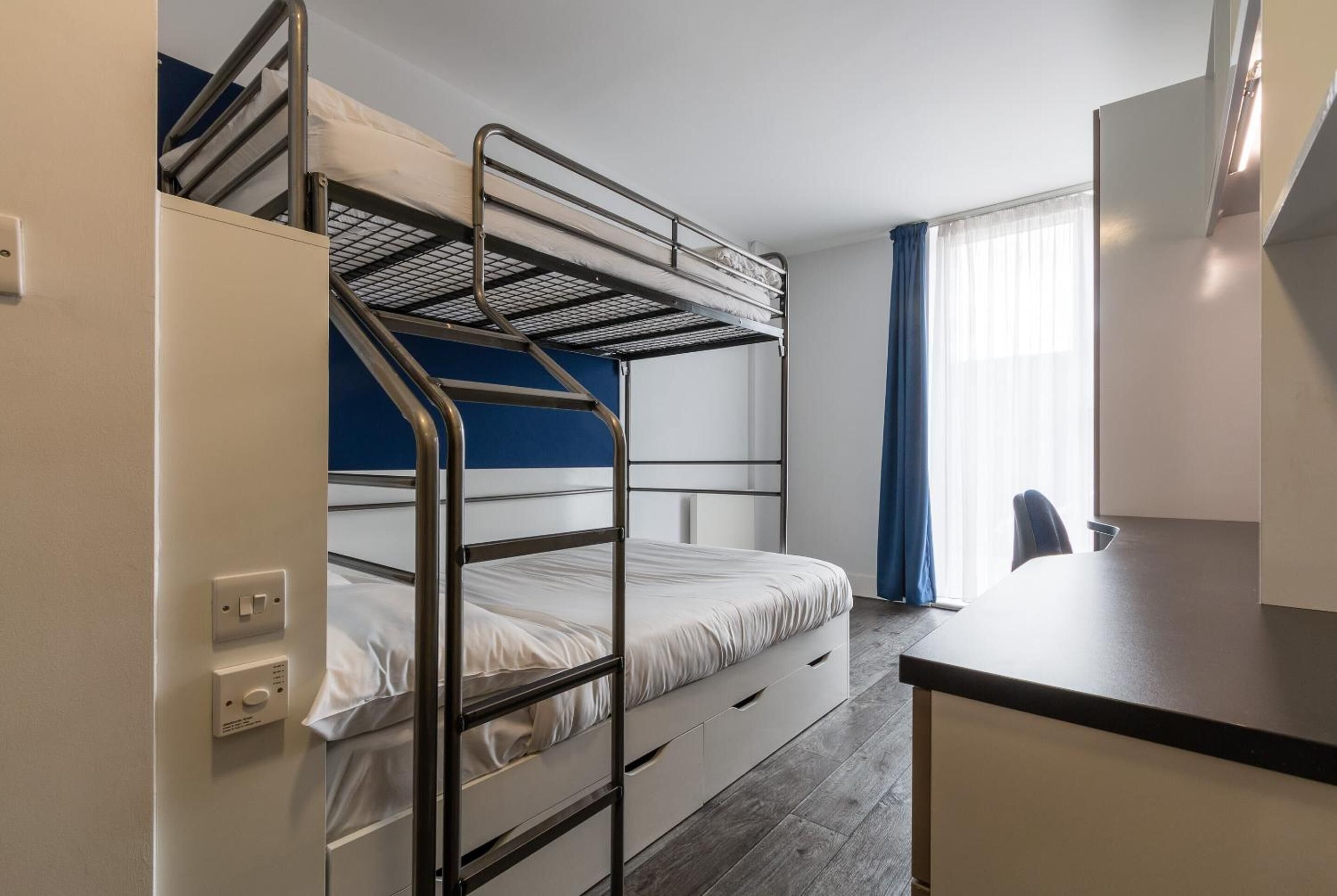 A bunk bed room with a desk and chair, a window with curtains, and a light switch on the wall.