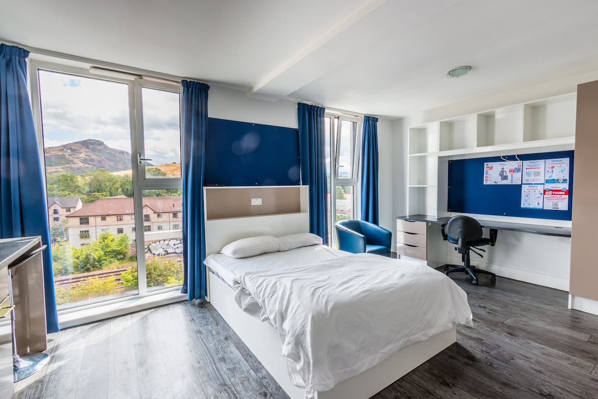 A modern hotel room with a large bed, a desk, and a view of the city through a large window.