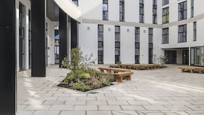 A modern building with a curved facade, multiple windows, and a courtyard with benches and plants. - Destiny Student Holyrood - Campus Accommodation (Edinburgh)