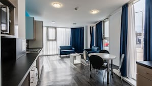 A modern hotel room with a kitchenette, a dining area with a table and chairs, a sofa, and large windows with curtains. - Destiny Student Holyrood - Campus Accommodation (Edinburgh)