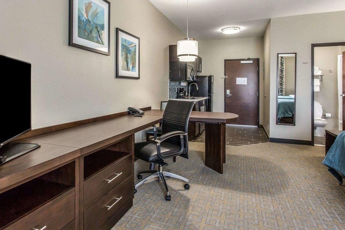 A hotel room with a desk, chair, television, artwork on the wall, and a bathroom visible through an open door.