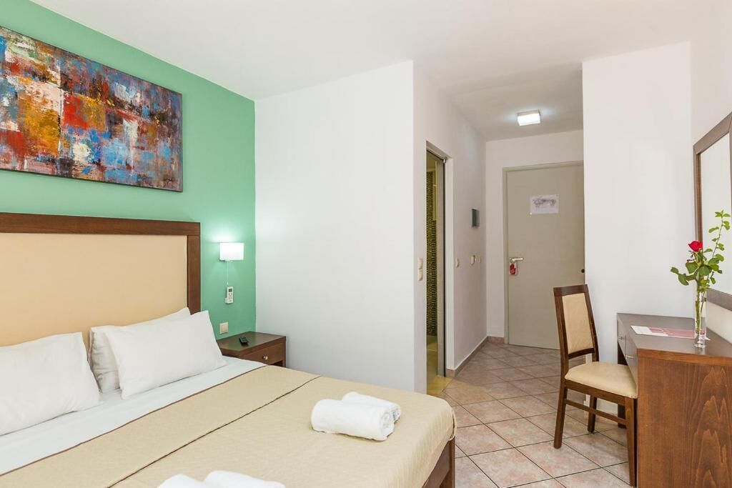 A hotel room with a bed, a desk, a chair, a painting on the wall, and a door leading to another room.