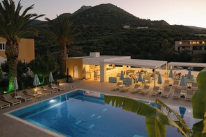 Une piscine avec des chaises longues et des parasols, un bâtiment comprenant un espace bar, et un paysage montagneux en arrière-plan.