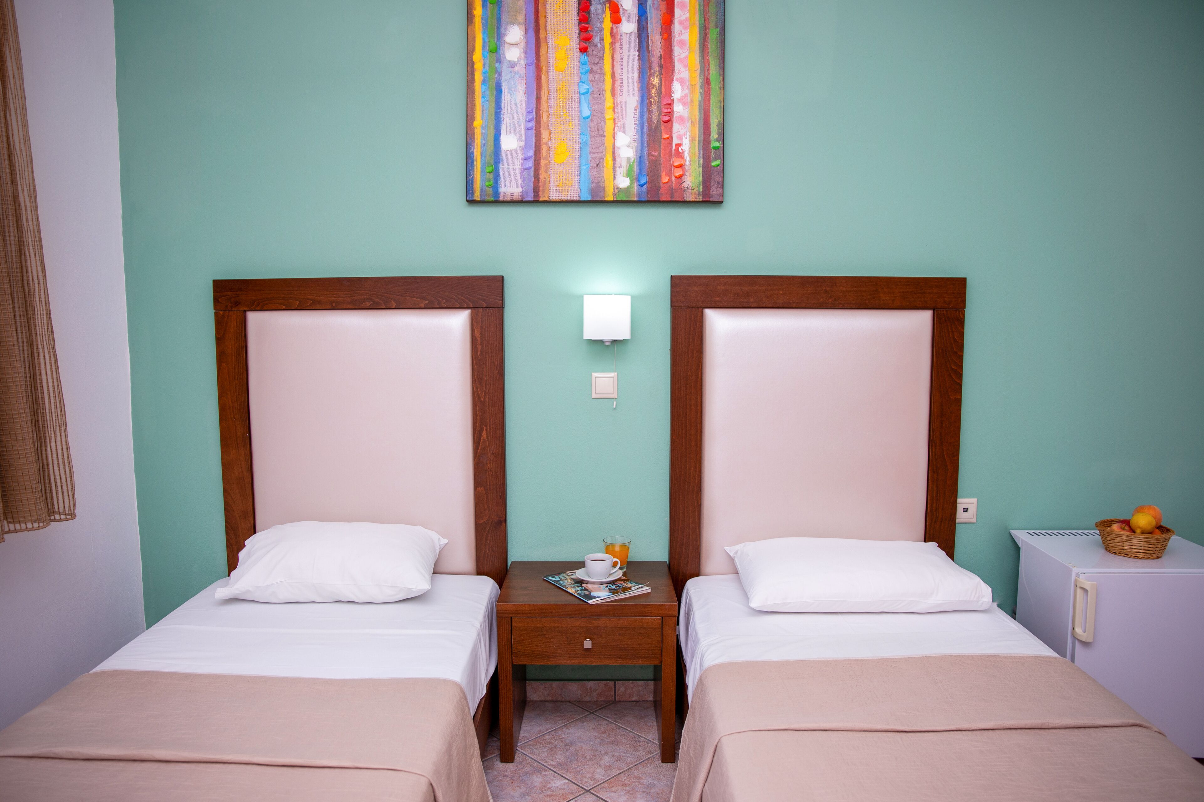 A hotel room with two single beds, a nightstand with a lamp, a small table with a basket of fruit, and a colorful abstract painting on the wall.