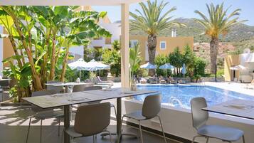An outdoor pool area with lounge chairs and umbrellas, surrounded by palm trees and buildings.