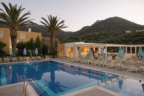 Une piscine avec des chaises longues et des parasols, un bâtiment doté d’une terrasse, et un paysage montagneux en arrière-plan.