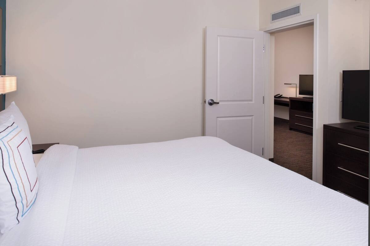 A hotel room with a bed, a desk with a computer, a television, and a door leading to another room.